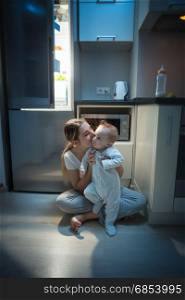 Young beautiful mother sitting with her baby son on kitchen floor at late night