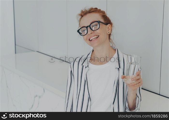 Young beautiful pleased red-haired woman having healthy habits, drinking mineral water in morning while standing in kitchen at home, smiling and enjoying new day, taking care of her body and skin. Young beautiful pleased red-haired woman having healthy habits, drinking mineral water in morning