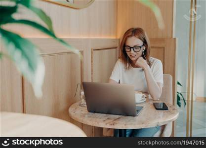 Young beautiful red-haired woman freelancer in earphones works remotely on laptop in cafe, female being concentrated at work while does remote job in cafeteria, enjoys cup of coffee in morning. Young beautiful red-haired woman freelancer in earphones works remotely on laptop in cafe