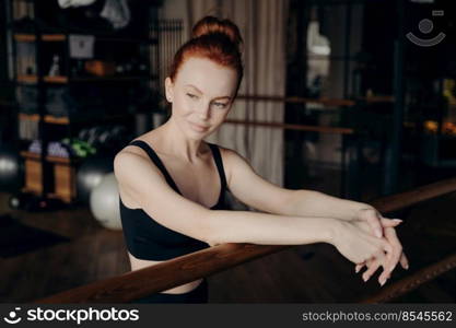 Young beautiful red haired woman with stylish bun leaning on ballet barre in fitness studio with blurred background while smiling and looking into distance, ballerina during stretching workout. Pretty young red haired woman leaning on ballet barre