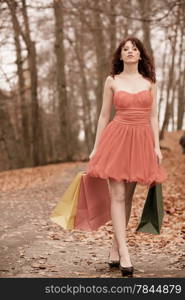 Young beautiful redhaired girl in elegant red dress with colorful shopping bags walking in autumnal park. Sale and retail concept. Outdoor.