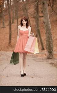 Young beautiful redhaired girl in elegant red dress with colorful shopping bags walking in autumnal park. Sale and retail concept. Outdoor.