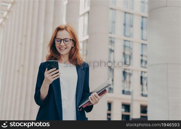 Young beautiful redhead female ceo executive looking at smartphone with excited face expression, reading news, browsing internet while going at business meeting with client, standing on city street. Young beautiful redhead female ceo executive looking at smartphone with excited face expression