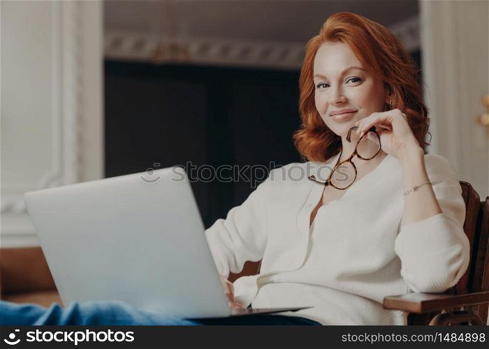 Young beautiful redhead skilled woman sends file to customer via modern laptop, sits in comfortable armchair, holds eyewear, spends time on online education, watches webinars, has pleased expession