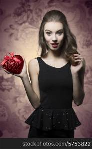 Young, beautiful, romantic, elegant, brunette girl in black dress and nice hairstyle is holding valentines heart. Her makeup is perfect.