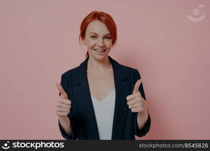 Young beautiful satisfied woman with red hair showing approval gesture thumbs up with both hands, positive female saying that everything is fine, dressed in black jacket, posing against pink wall. Young beautiful satisfied woman with red hair showing approval gesture thumbs up with both hands