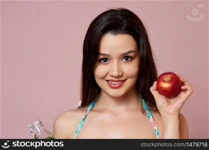 young beautiful slender Asian girl in a swimsuit . with fresh juice and an Apple in his hands. smiles. on a pink isolated background.