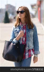 Young beautiful stylish girl in sunglasses and with a bag in hand on a sunny day on the streets of major cities