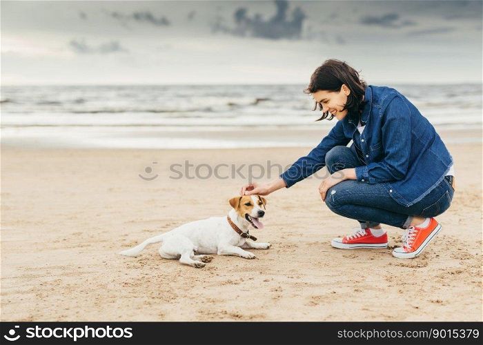 Young beautiful stylish woman flatters dog on seashore, enjoys spending weekends outdoor with pet, being fond of animals. Female glad to have walk with dog. Lifestyle and leisure concept. Woman flatters dog.
