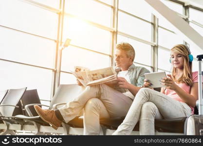 Young beautiful teenage girl watching movie on digital tablet with headphones on while her father is reading newspaper in airport