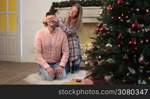 Young beautiful wife keeping her husband&acute;s eyes covered with hands while cute daughter giving Christmas gift in xmas decorated room. Adorable little girl giving present to her handsome father during family winter holidays at home. Dolly shot.