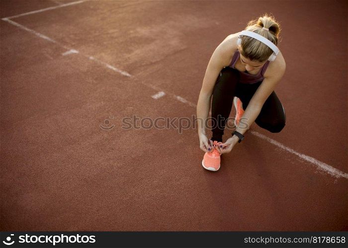Young beautiful woman athlete with  earphones listening to music and tying laces during training in park with sunset and sunbeam on background