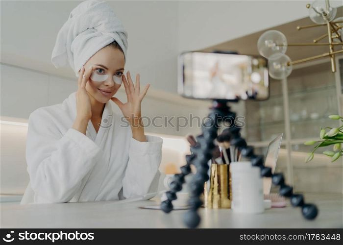 Young beautiful woman blogger in bathrobe recording video tutorial for beauty blog or social media how to use cosmetic patches or pads from dark circles while sitting in kitchen. Skin care concept. Female beauty blogger in bathrobe using cosmetic patches while recording video about skin care for her vlog