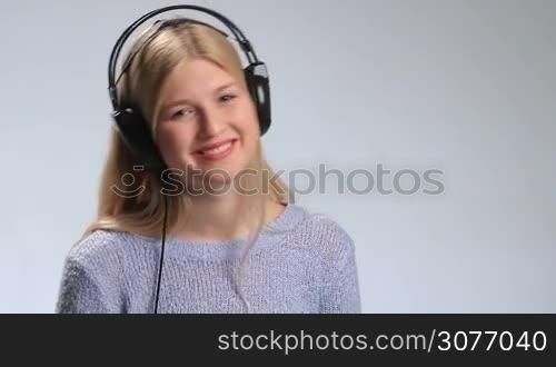Young beautiful woman enjoying music with headphones on white. Cheerful teenage girl in black headphones touching the earcups with hand, swaying her head to the rhythm of song and smiling into the camera