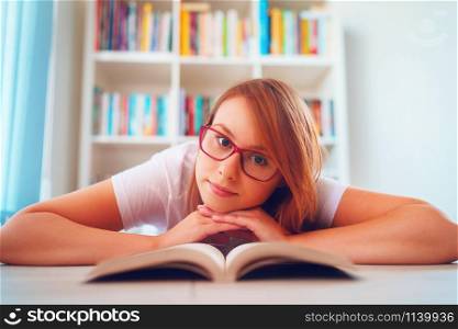 Young beautiful woman girl student lying on the floor in front of the book shelves at home study or read with her head over hands wearing glasses looking to the camera