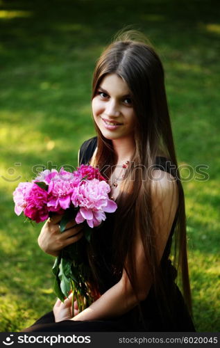 Young beautiful woman in a black dress posing with a bouquet of peonies in a garden. Fashion model girl with bunch of flowers outdoors in summer