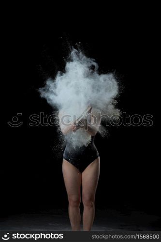 young beautiful woman in a black textile bodysuit holds a book in her hands, cloud of white wheat flour flies out of the book in different directions, explosion. Sportswoman stands on black background