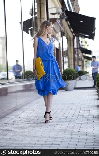 Young beautiful woman in a blue dress and a yellow bag on the background of shop windows