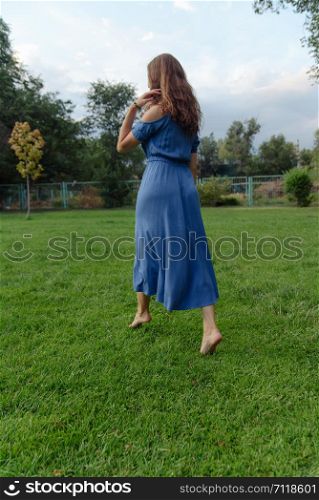 Young beautiful woman in a blue dress walks barefoot on the grass in the park and smiles. A young woman in a blue dress walks barefoot on the grass in the park