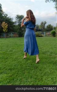Young beautiful woman in a blue dress walks barefoot on the grass in the park and smiles. A young woman in a blue dress walks barefoot on the grass in the park