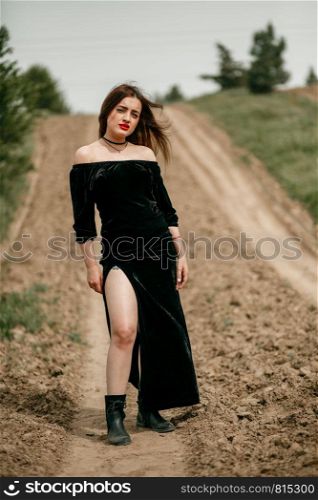 Young beautiful woman in black dress posing for a photographer on a sandy road.