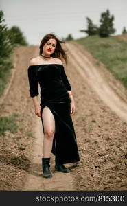 Young beautiful woman in black dress posing for a photographer on a sandy road.