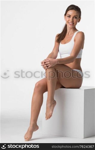 Young beautiful woman in cotton underwear sitting and touching own legs skin, white background. Woman in underwear touch legs