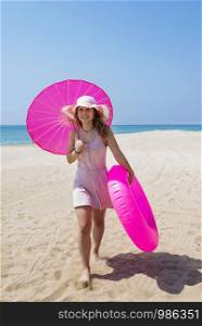 Young beautiful woman in pink dress walking on sand beach wearing a hat and holding an umbrella and a floater