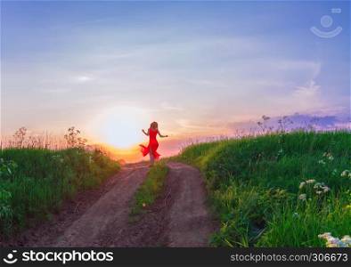 Young beautiful woman in red dress is dancing in the distance on a dirt path in a green field against the setting sun in the evening sky.. Young Woman Dancing Against The Sunset