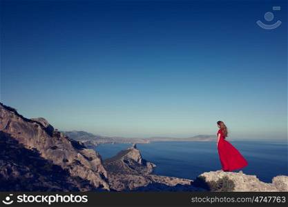 Young beautiful woman in red dress looking to mountains sea. Girl on the nature on blue sky background. Fashion photo