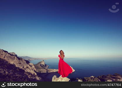 Young beautiful woman in red dress looking to mountains sea. Girl on the nature on blue sky background. Fashion photo