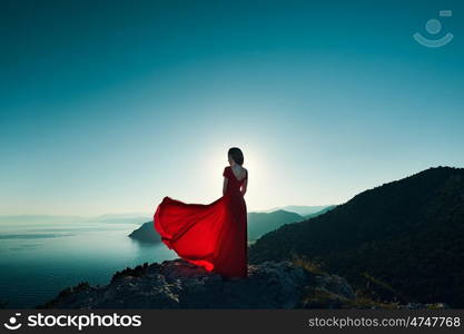 Young beautiful woman in red dress looking to mountains sea. Girl on the nature on blue sky background. Fashion photo