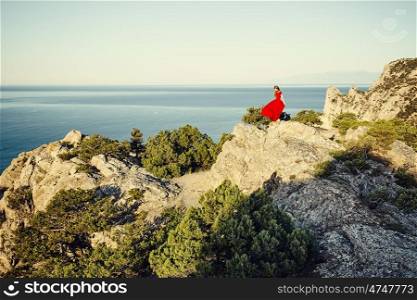 Young beautiful woman in red dress looking to mountains sea. Girl on the nature on blue sky background. Fashion photo