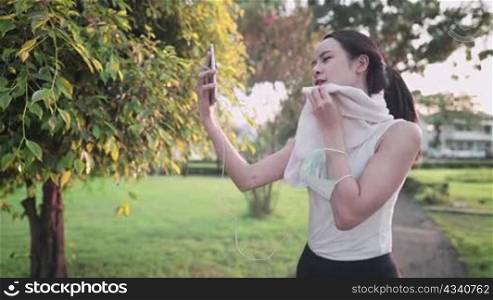 Young beautiful woman in sport clothing wiping sweat from face, video calling through smartphone during exercising at the park, rest after exercise sharing healthy lifestyle, get online with friends