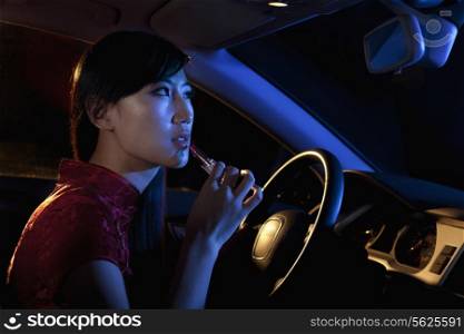 Young beautiful woman in traditional Chinese dress putting on lipstick in the rear view mirror of the car at night