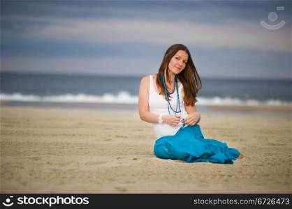 Young beautiful woman in white cami and turquoise skirt sits on sand near the ocean