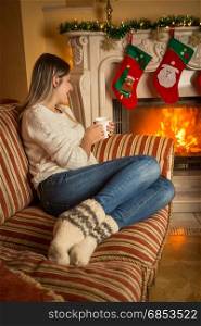 Young beautiful woman in woolen socks relaxing on sofa at burning fireplace and drinking tea