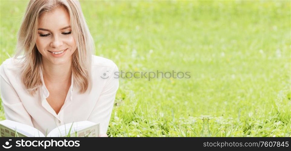 Young beautiful woman lays on green grass field and reads book, spring exam concept, copy space for text. Woman lays on grass and reads book