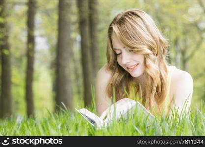 Young beautiful woman lays on green grass field and reads book, spring exam concept, copy space for text. Woman lays on grass and reads book