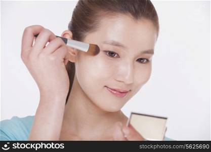 Young beautiful woman looking into a mirror and applying make up with a make up brush, studio shot