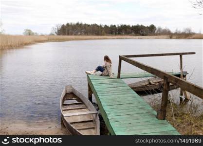 Young beautiful woman sits on the wooden bridge on the river at spring day. old boat near the shore. selective focus. Young beautiful woman sits on the wooden bridge on the river at spring day. old boat near the shore. selective focus.