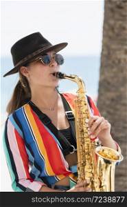 Young beautiful woman wearing a hat and glasses playing a saxophone on an out of focus background. Street performer and lifestyle concept.