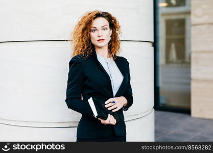 Young beautiful woman wearing formal clothes, dressed formally, holding pocketbook with pen, having bushy hairstyle, standing near office builduing. Successful pretty female office worker working