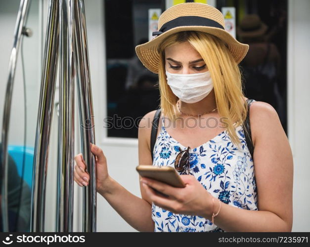 Young beautiful woman wears face mask and keeps social distance by standing and using smart phone while transporting at train during covid-19 or coronavirus outbreak. Young beautiful woman keeps social distance during covid19