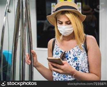 Young beautiful woman wears face mask and keeps social distance by standing and using smart phone while transporting at train during covid-19 or coronavirus outbreak. Young beautiful woman keeps social distance during covid19