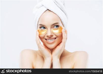 Young beautiful woman with healthy fresh skin, applies golden patches under eyes against dark circles, shows effect of perfect cosmetic product, stands shirtless, isolated on white background.
