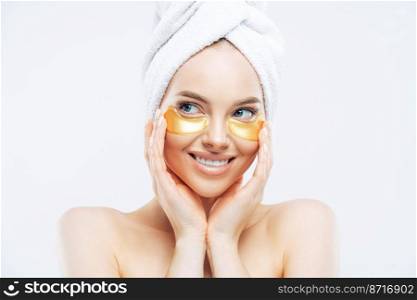 Young beautiful woman with healthy fresh skin, applies golden patches under eyes against dark circles, shows effect of perfect cosmetic product, stands shirtless, isolated on white background.