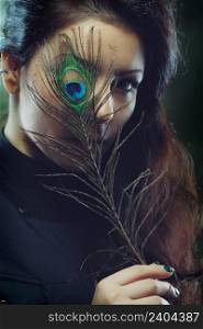 Young beautiful woman with long curly hair holding peacock feather in front of her face, dark green background