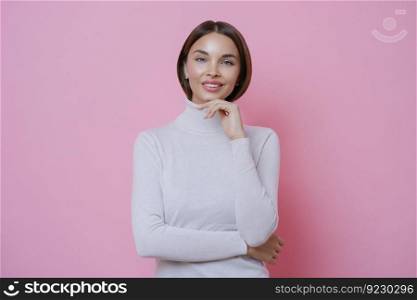 Young beautiful woman with pleased expression, touches chin, smiles tenderly, has straight dark hair, dressed in casual white turtleneck, isolated on pink background. Femininity, beauty and skin care