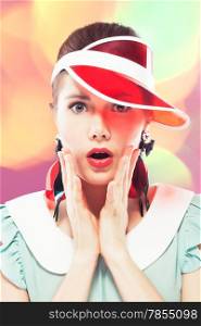 Young beautiful woman with red glamour lips and eye arrow make-up wearing fancy plastic earrings and red sun visor on multicolor background, she is open-mouthed with surprise, retro beauty style